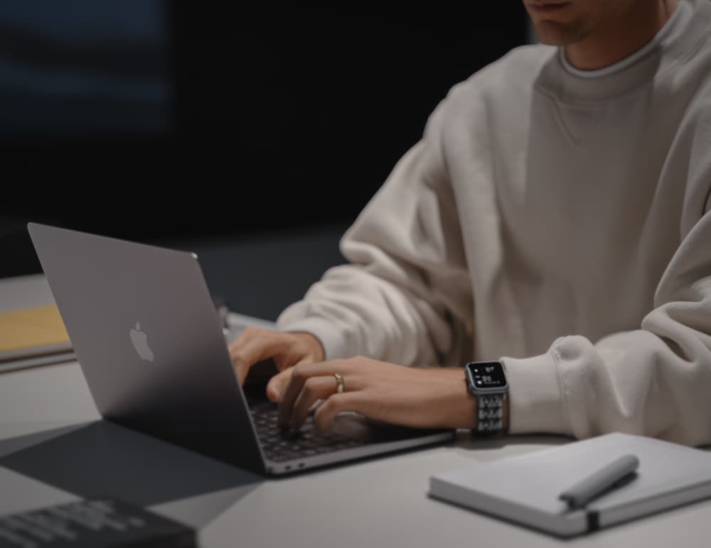 A man in a cream sweatshirt sitting at a laptop working with other things on his desk